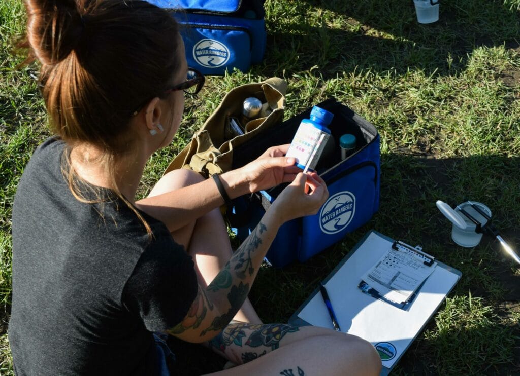 A woman tests chlorine in freshwater using a Water Rangers testkit. She is comparing a test strip to the reference scale on the back of the bottle.